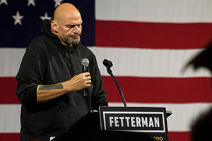 Senator John Fetterman holding a mic and standing behind a podium and before an American flag.
